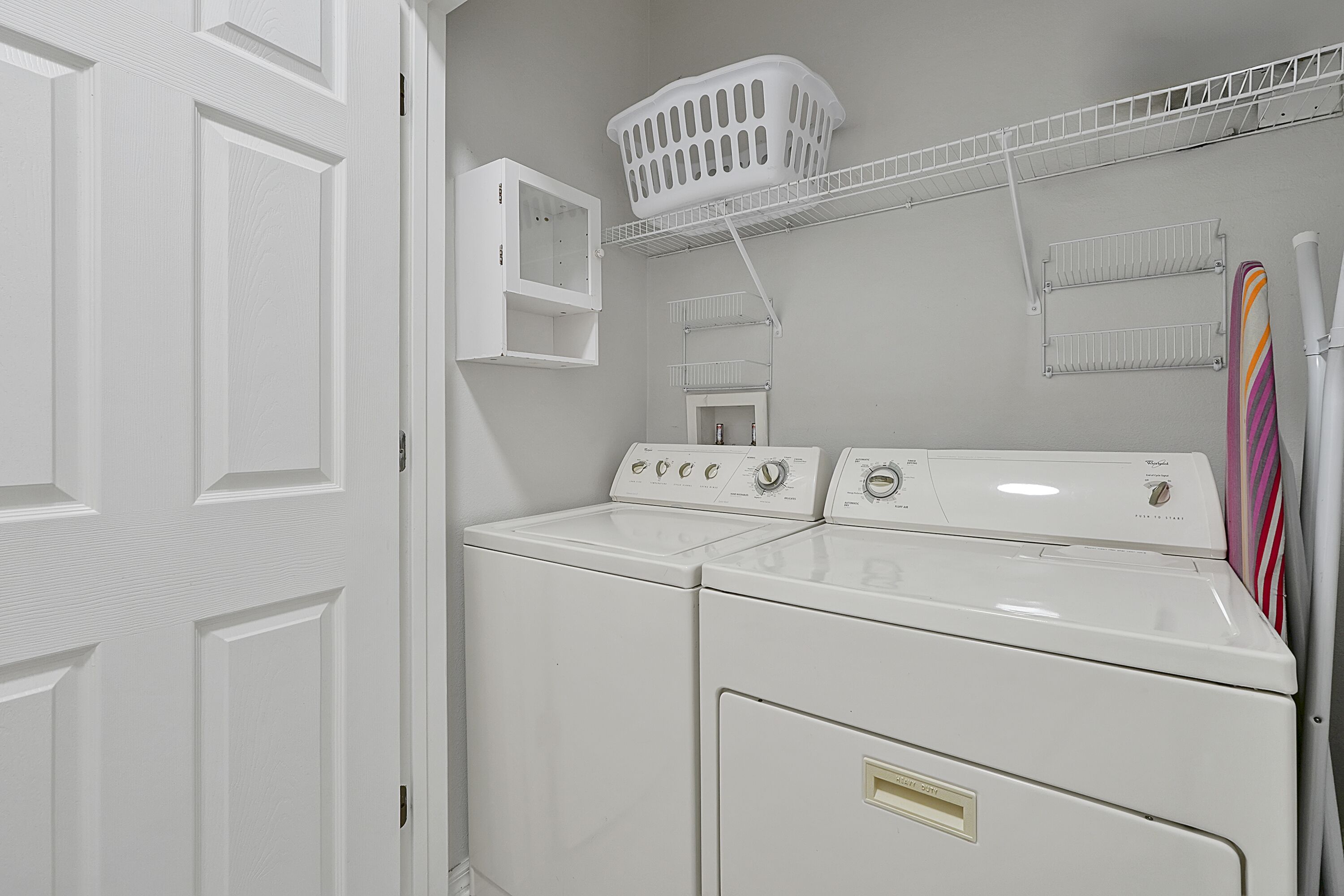 A laundry room with two white front-loading washers and dryers, a white laundry basket on a shelf, and a striped towel hanging on the wall.