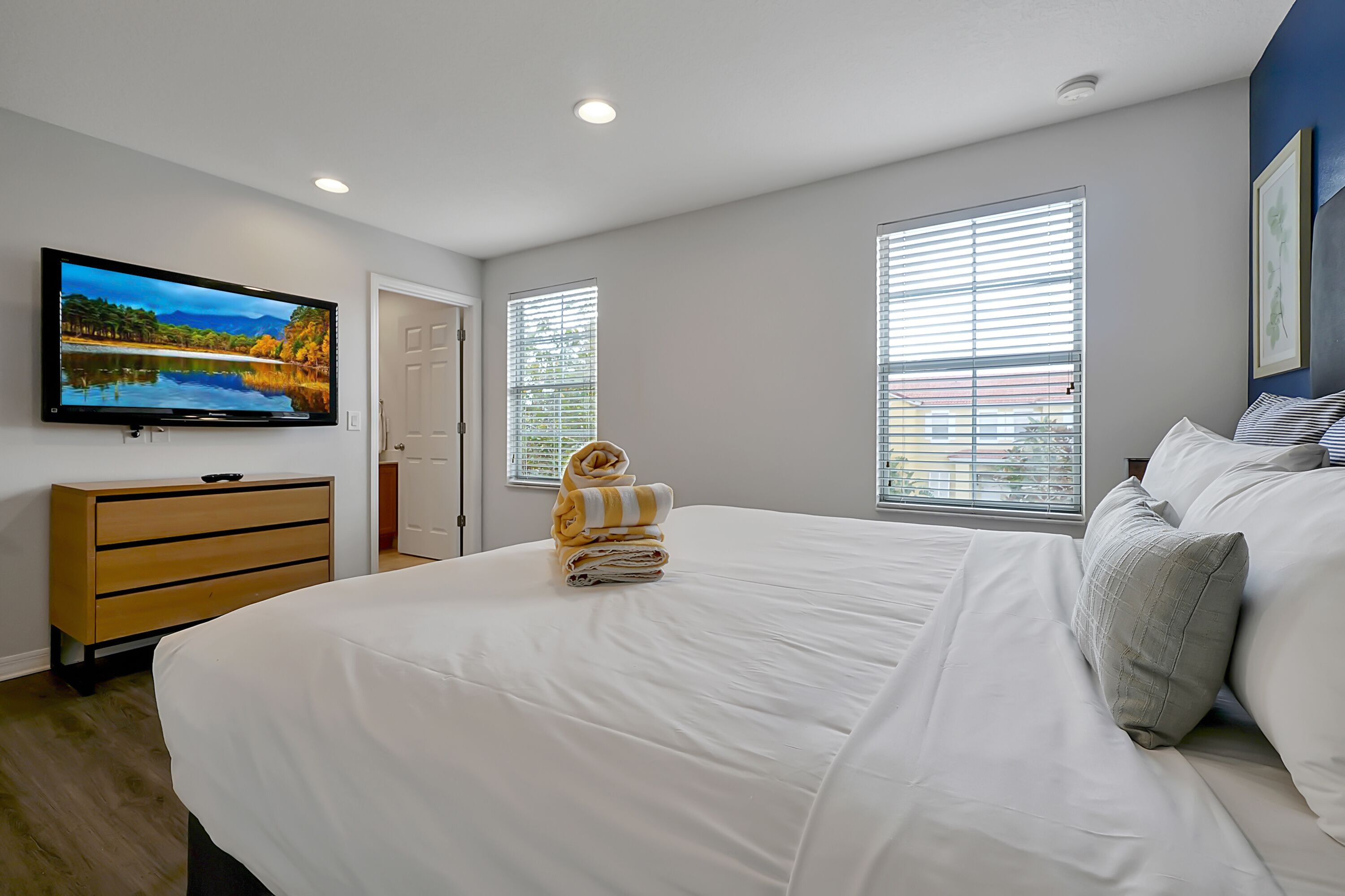 Un dormitorio con una cama grande, un televisor en la pared y dos ventanas con persianas.