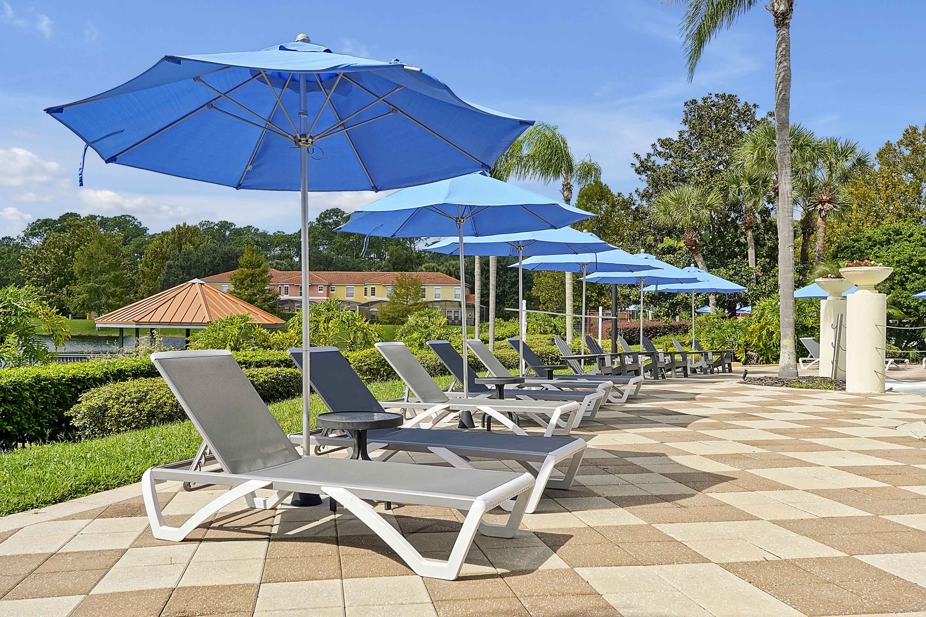A series of sun loungers with blue umbrellas, arranged in rows on a checkered tile patio.