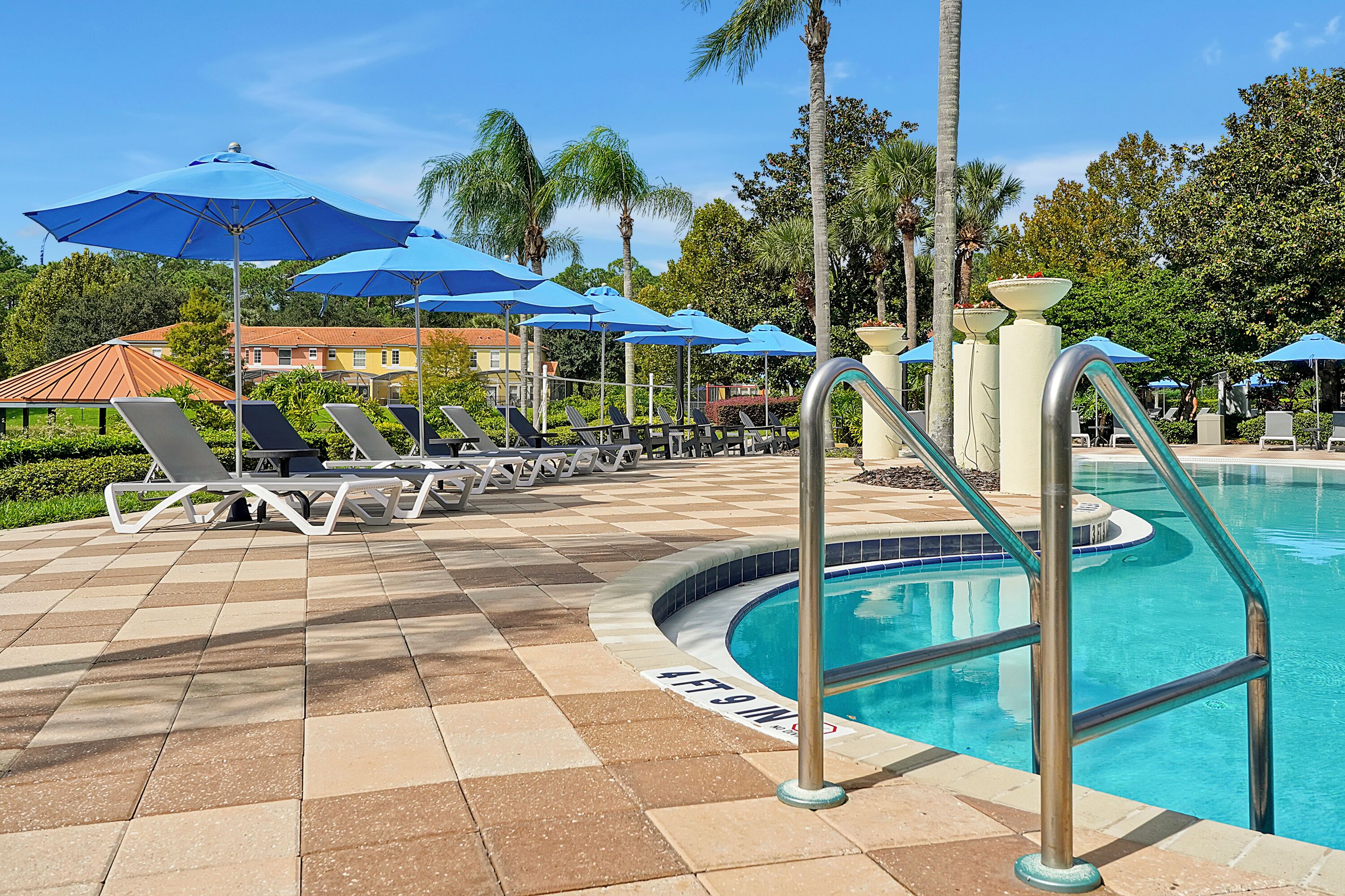 Zona junto a la piscina con sillones y sombrillas, una piscina con borde de azulejos y palmeras al fondo.