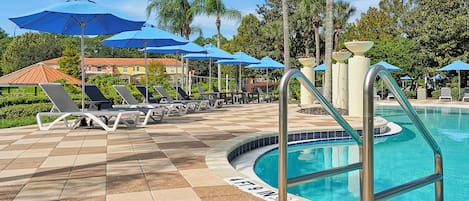 Zona junto a la piscina con sillones y sombrillas, una piscina con borde de azulejos y palmeras al fondo.