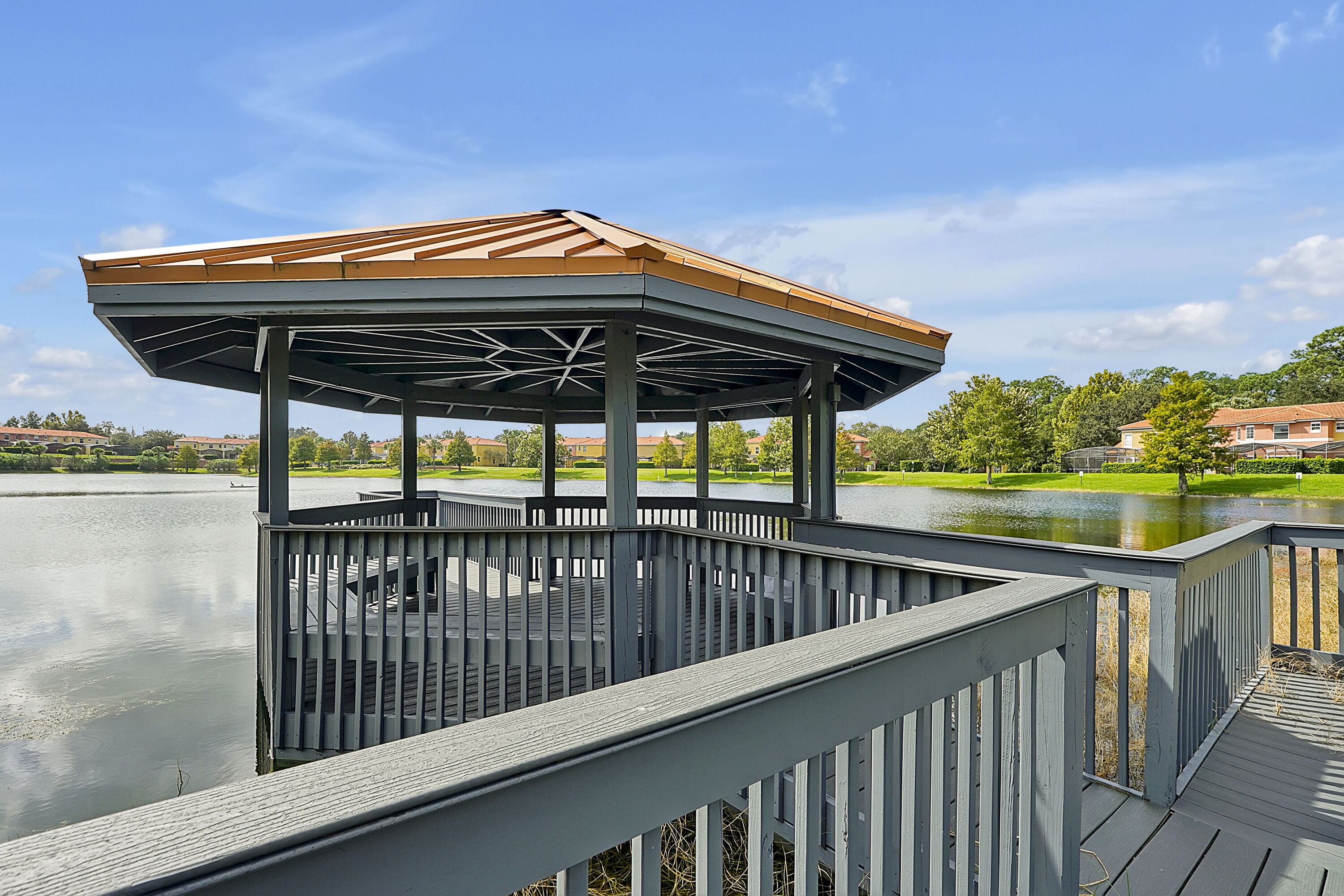 A gazebo with a roof and railings overlooking a body of water, surrounded by greenery and buildings.