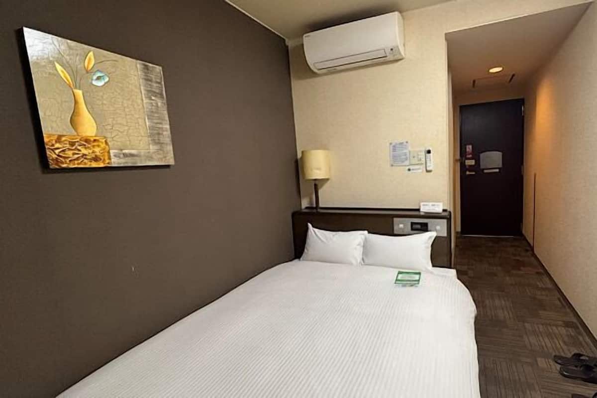 A hotel room with a bed, a wall-mounted air conditioner, a painting, a lamp, and a door leading to another room.