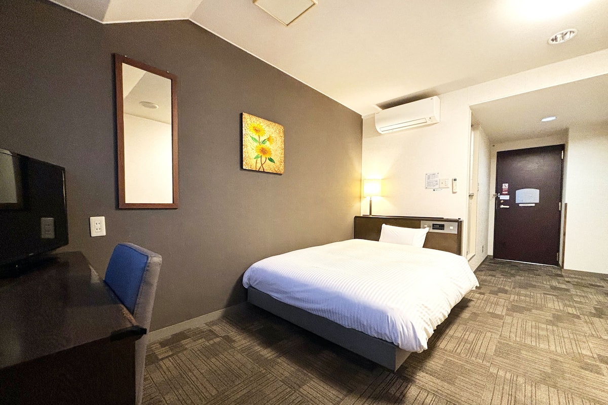 A hotel room with a bed, a desk, a chair, a mirror, a painting, a lamp, and a door.