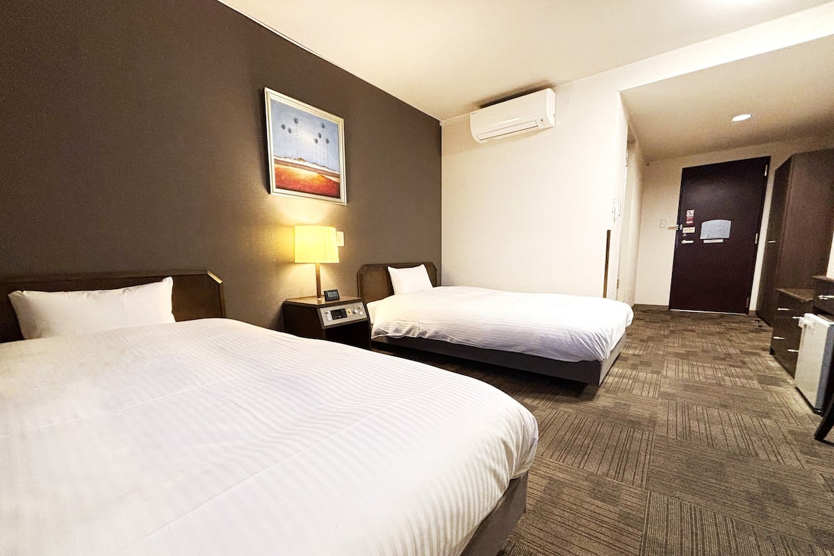 A hotel room with two beds, a nightstand with a lamp, an air conditioning unit, and a door to the hallway.