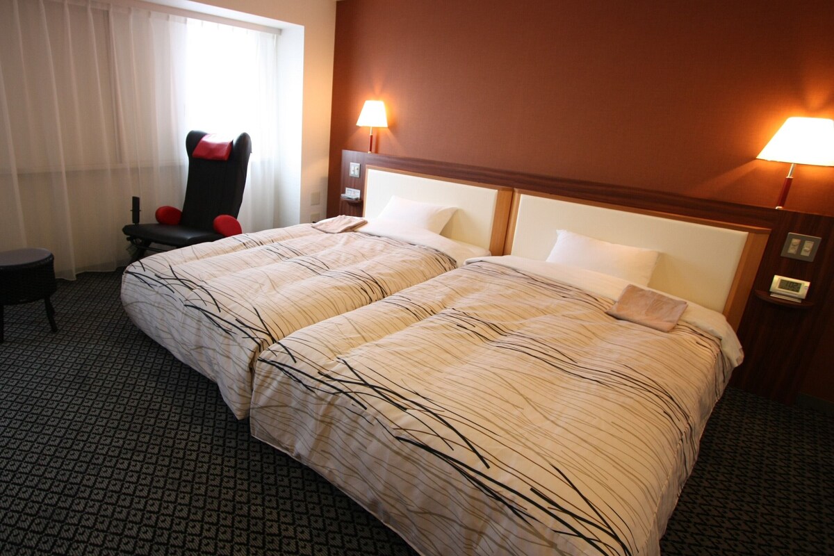 A hotel room with a large bed, two bedside lamps, a chair with red cushions, a small table, and a window with sheer curtains.