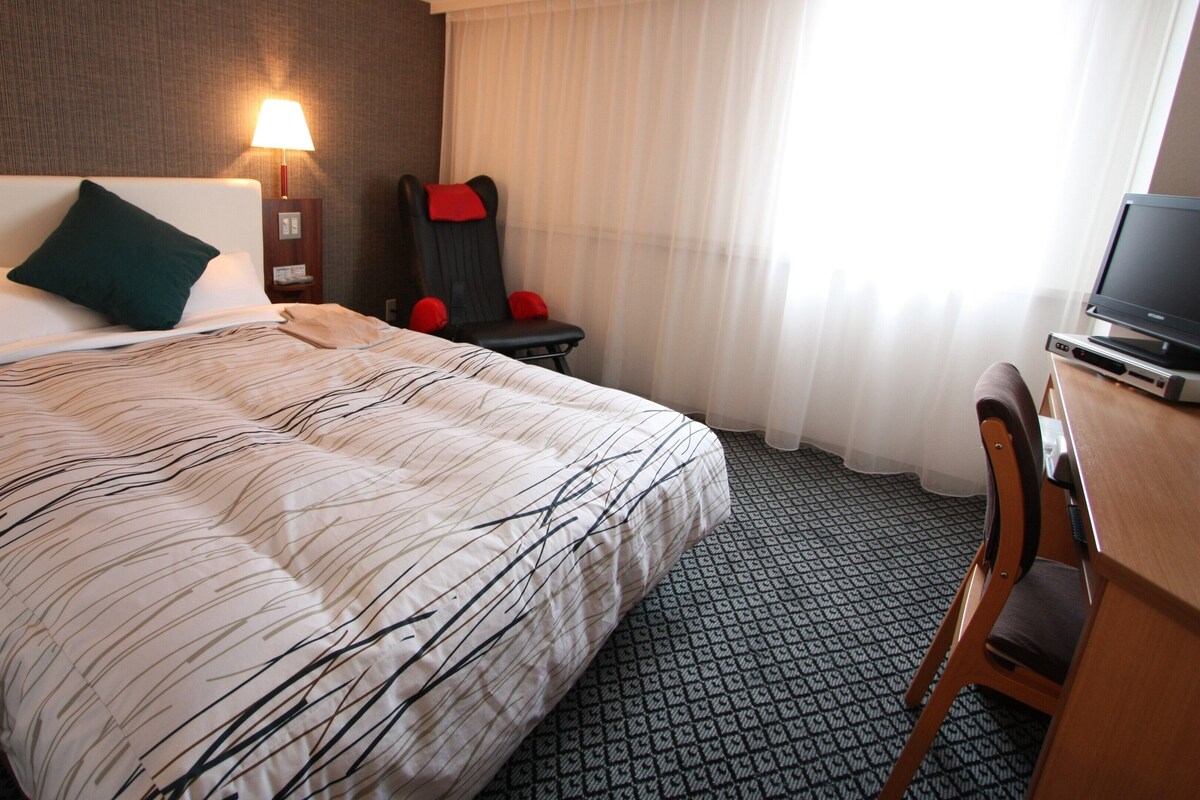A hotel room with a bed, a desk, a chair, a TV, and a window with curtains.