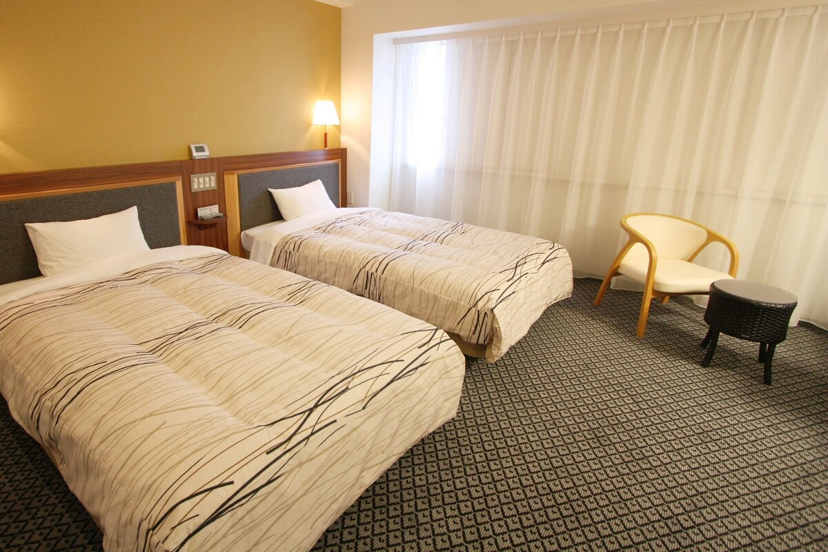 A hotel room with two beds, a chair, a small table, and a window with curtains.