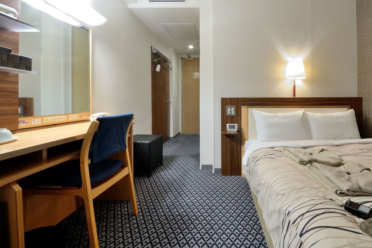 A hotel room with a bed, a desk with a chair, a mirror, a wall-mounted lamp, and a clock.