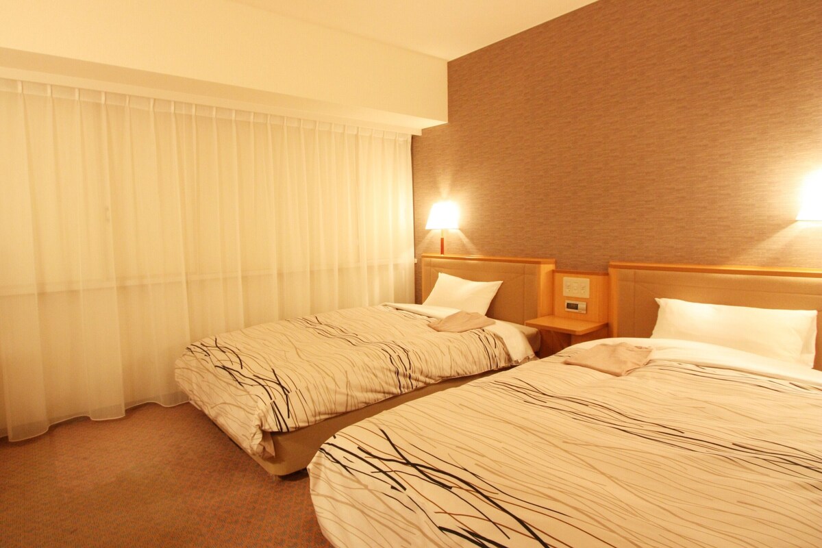 A hotel room with two beds, a TV, a desk, and a window with curtains.