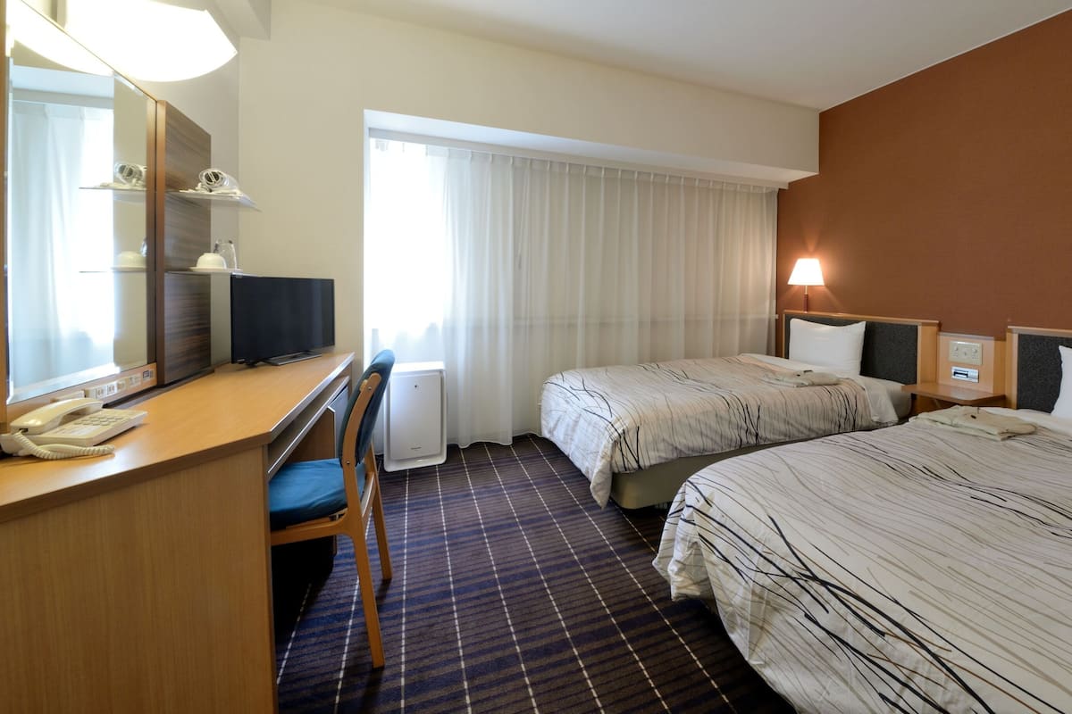 A hotel room with a desk, chair, two beds, a mirror, a lamp, and a window with curtains.