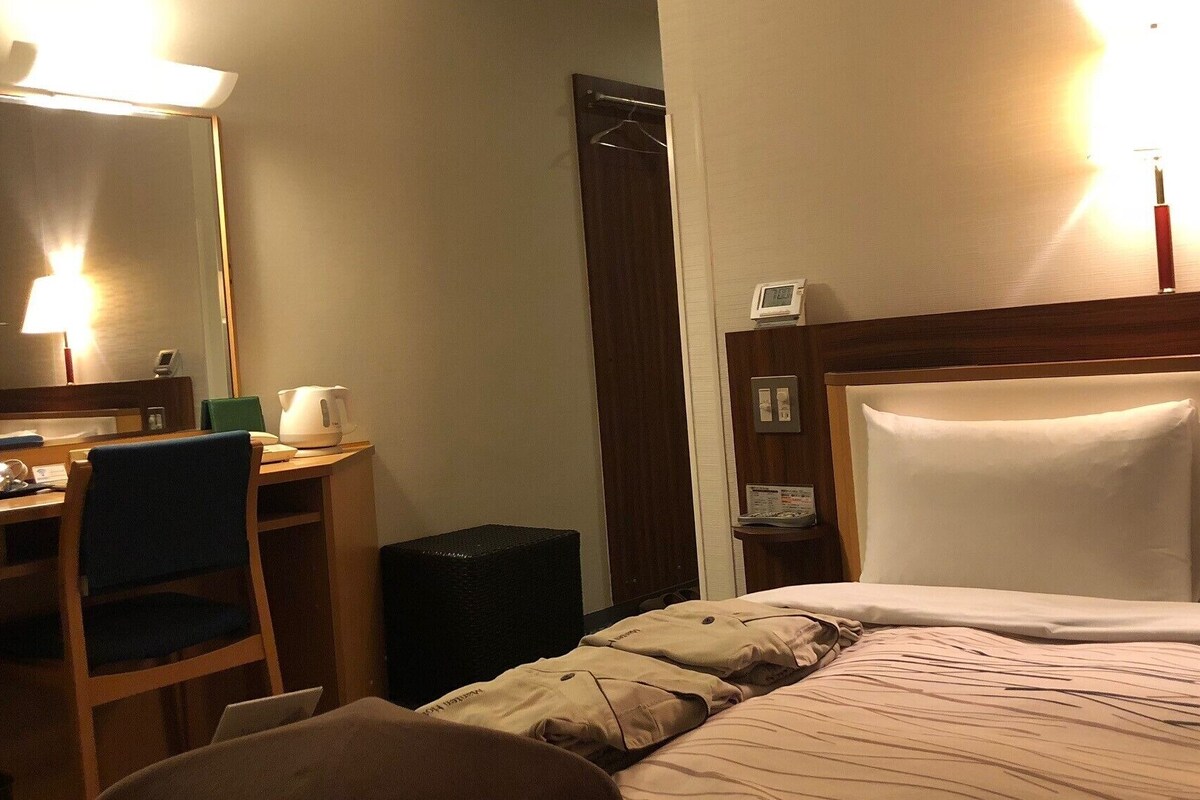 A hotel room with a bed, a desk with a chair, a mirror, and a bedside table with a lamp.