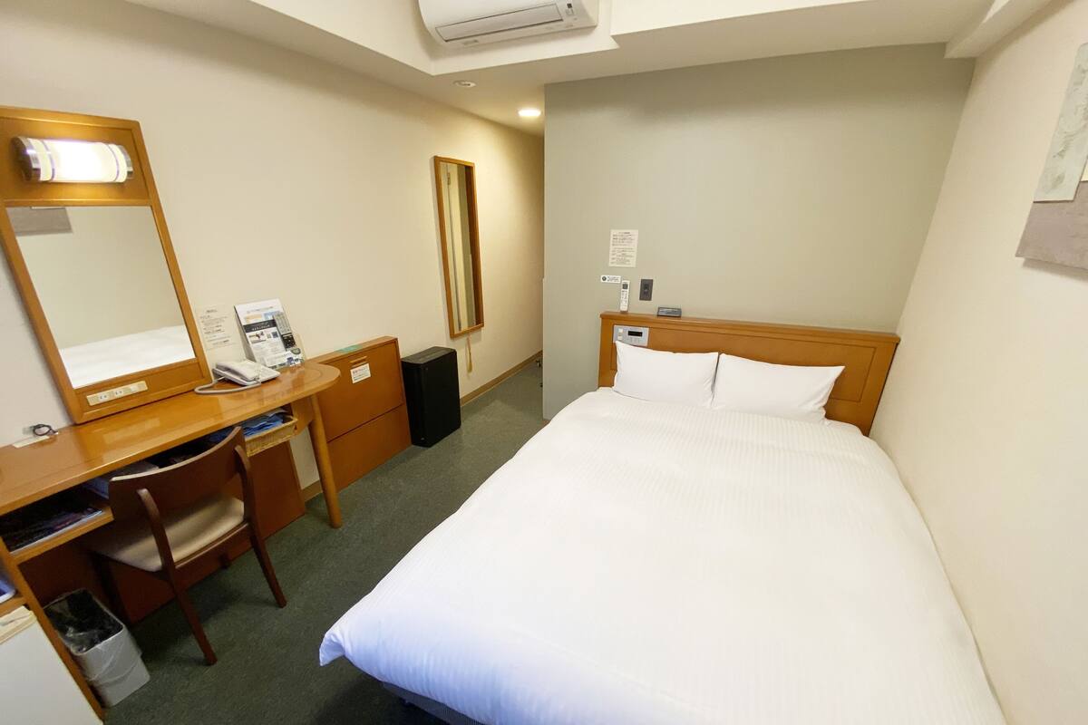A hotel room with a bed, a desk, a chair, a mirror, a telephone, and a small trash bin.
