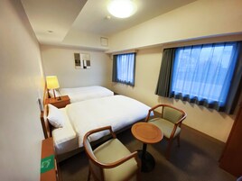 Habitación de hotel con dos camas, una mesita, dos sillas, una lámpara y un ventanal con cortinas.