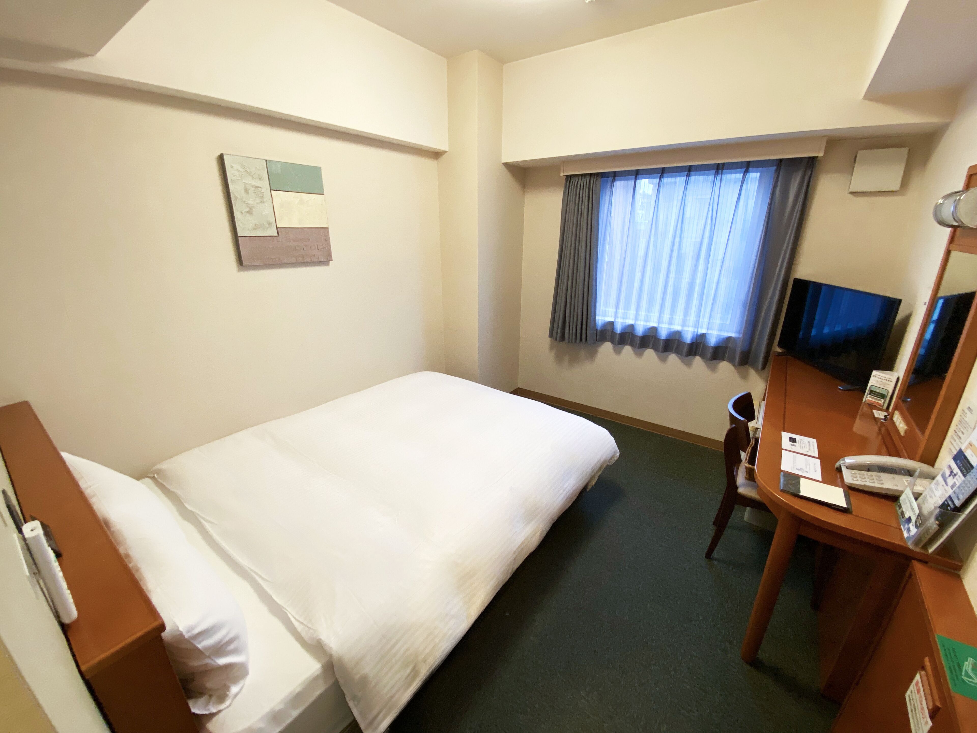 Una habitación de hotel con una cama individual, un escritorio con teléfono, un televisor y una ventana con cortinas.