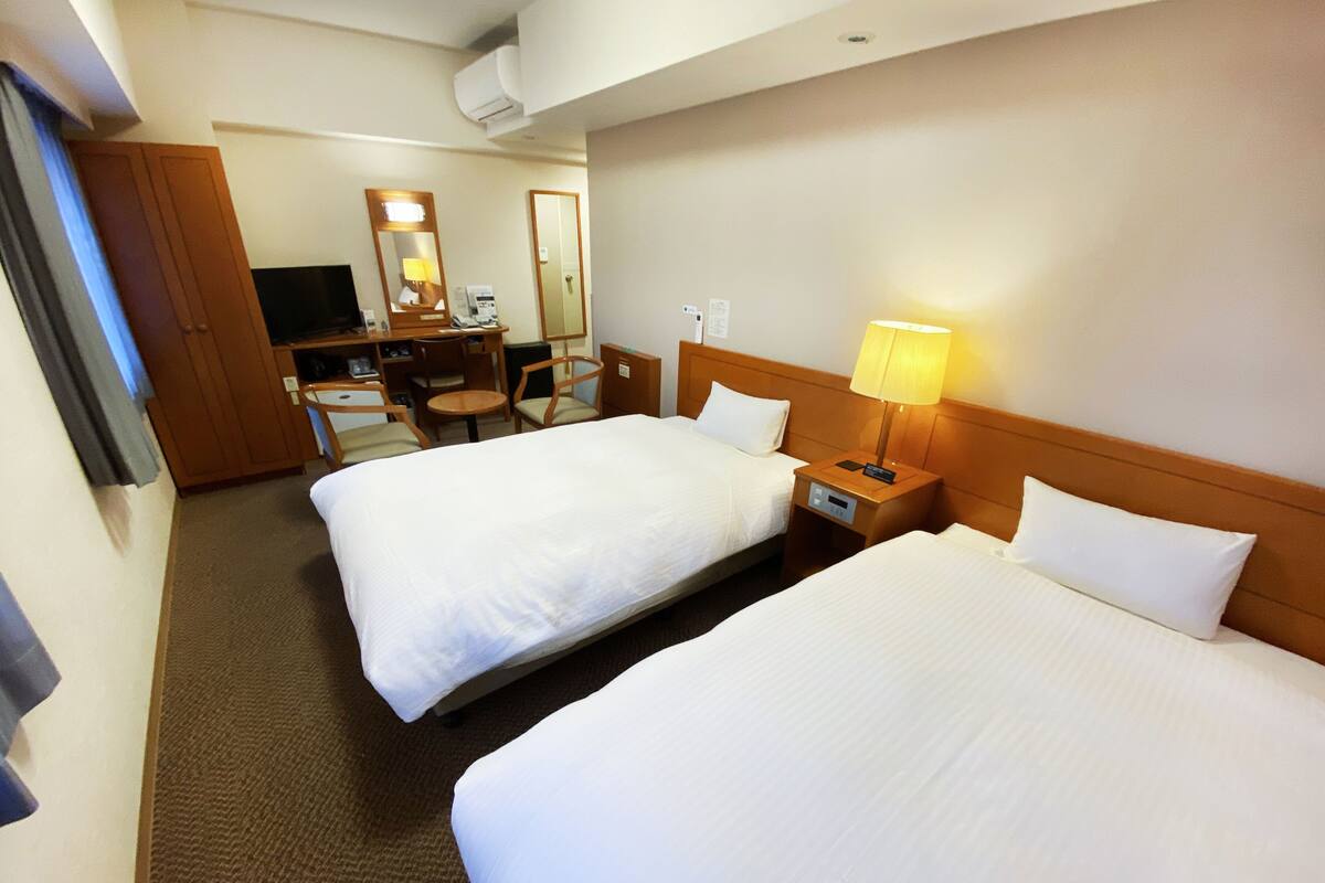 A hotel room with two beds, a desk with a chair, a lamp, and a TV.