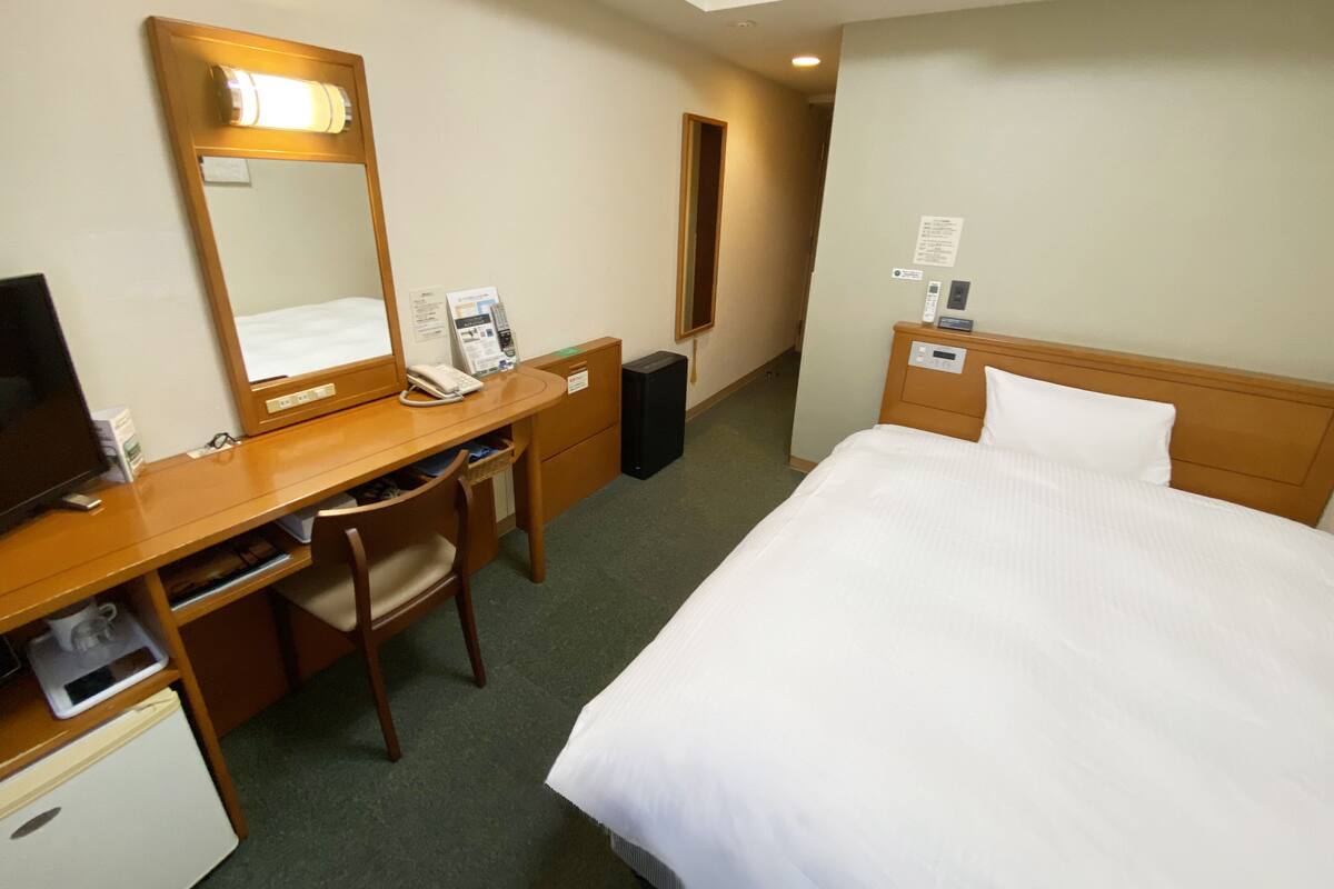 A hotel room with a bed, a desk with a chair, a mirror, a telephone, and a small refrigerator.