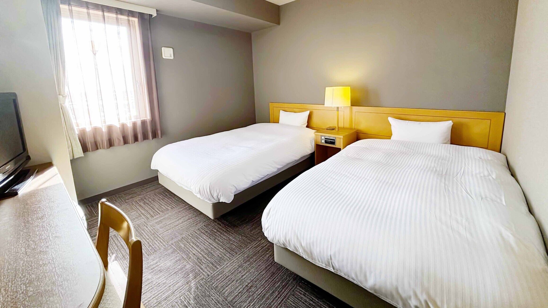 A hotel room with two beds, a desk, a chair, a TV, and a window with curtains.