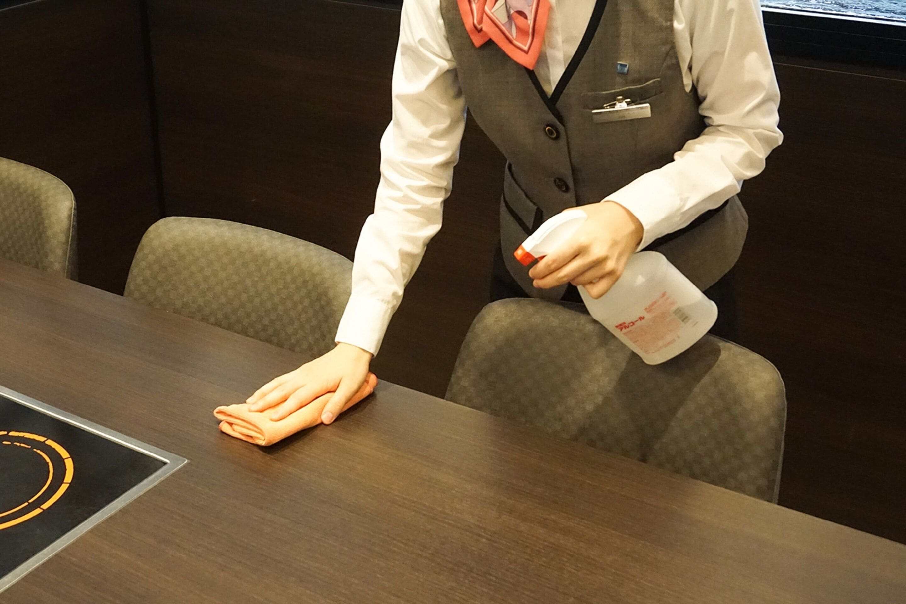 A person in a white shirt and grey vest cleaning a wooden table with a cloth and a bottle of cleaning solution.