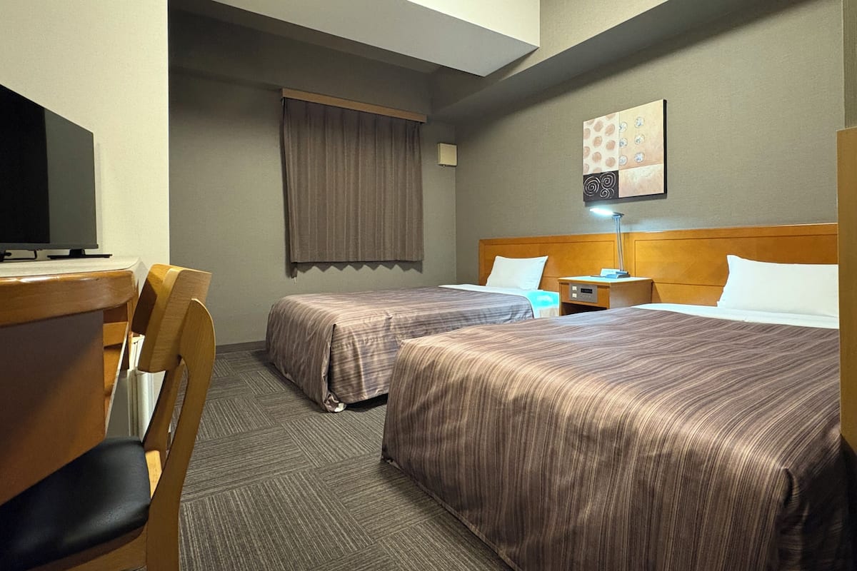 A hotel room with two beds, a desk, a chair, a TV, a lamp, and a painting on the wall.