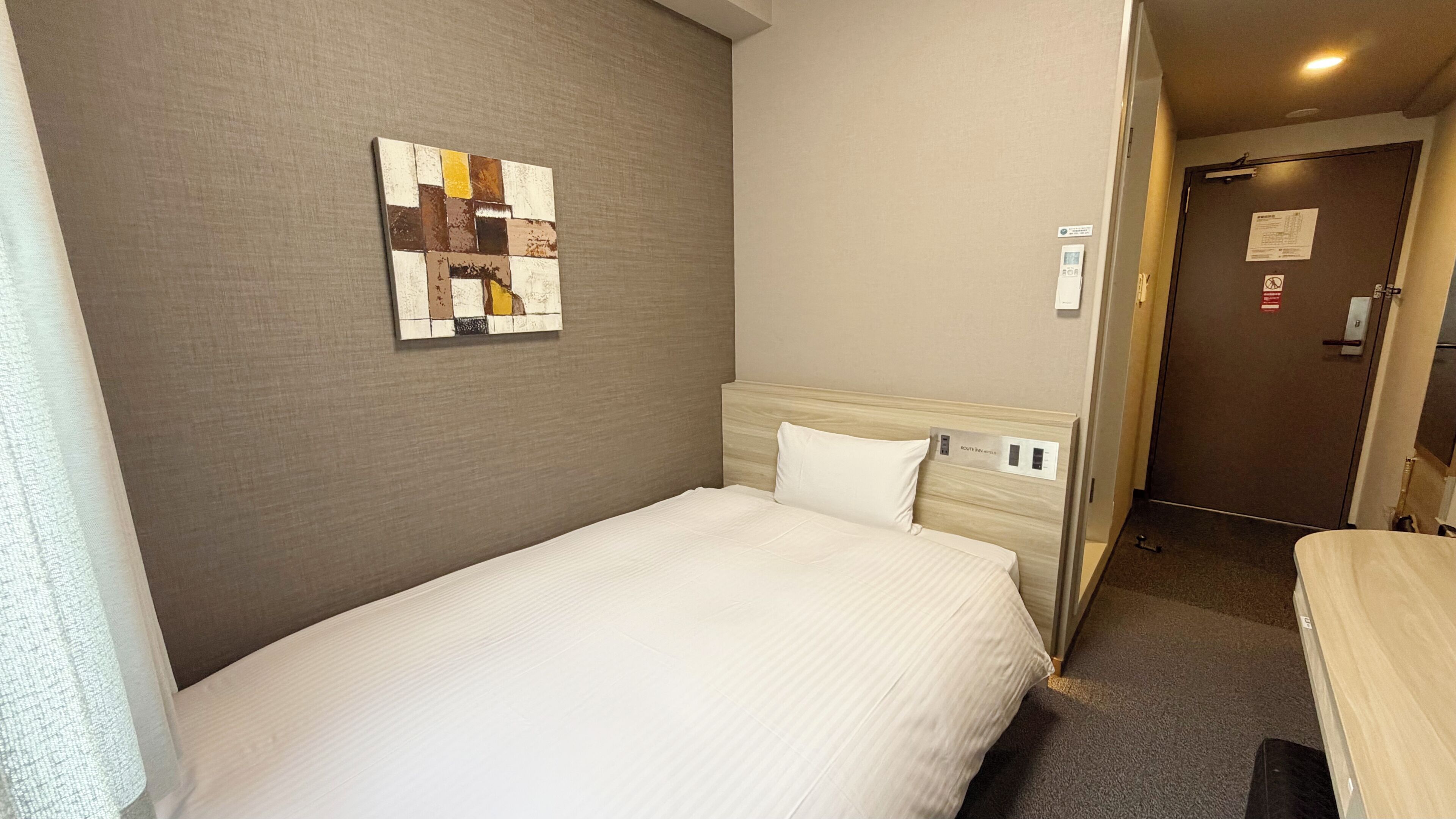 A hotel room with a single bed, a headboard with a control panel, a wall-mounted artwork, and a door with a sign.