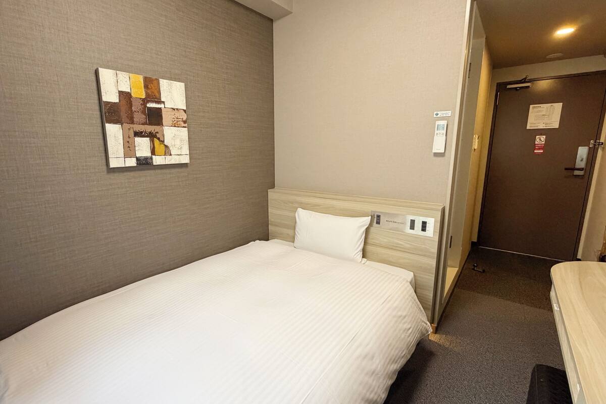 A hotel room with a single bed, a headboard with a control panel, a wall-mounted artwork, and a door with a sign.