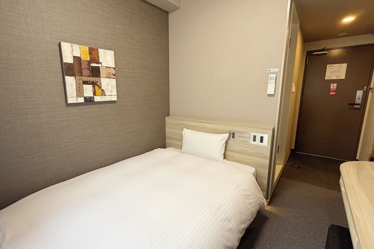 A hotel room with a single bed, a headboard with a control panel, a wall-mounted artwork, and a door with a sign.