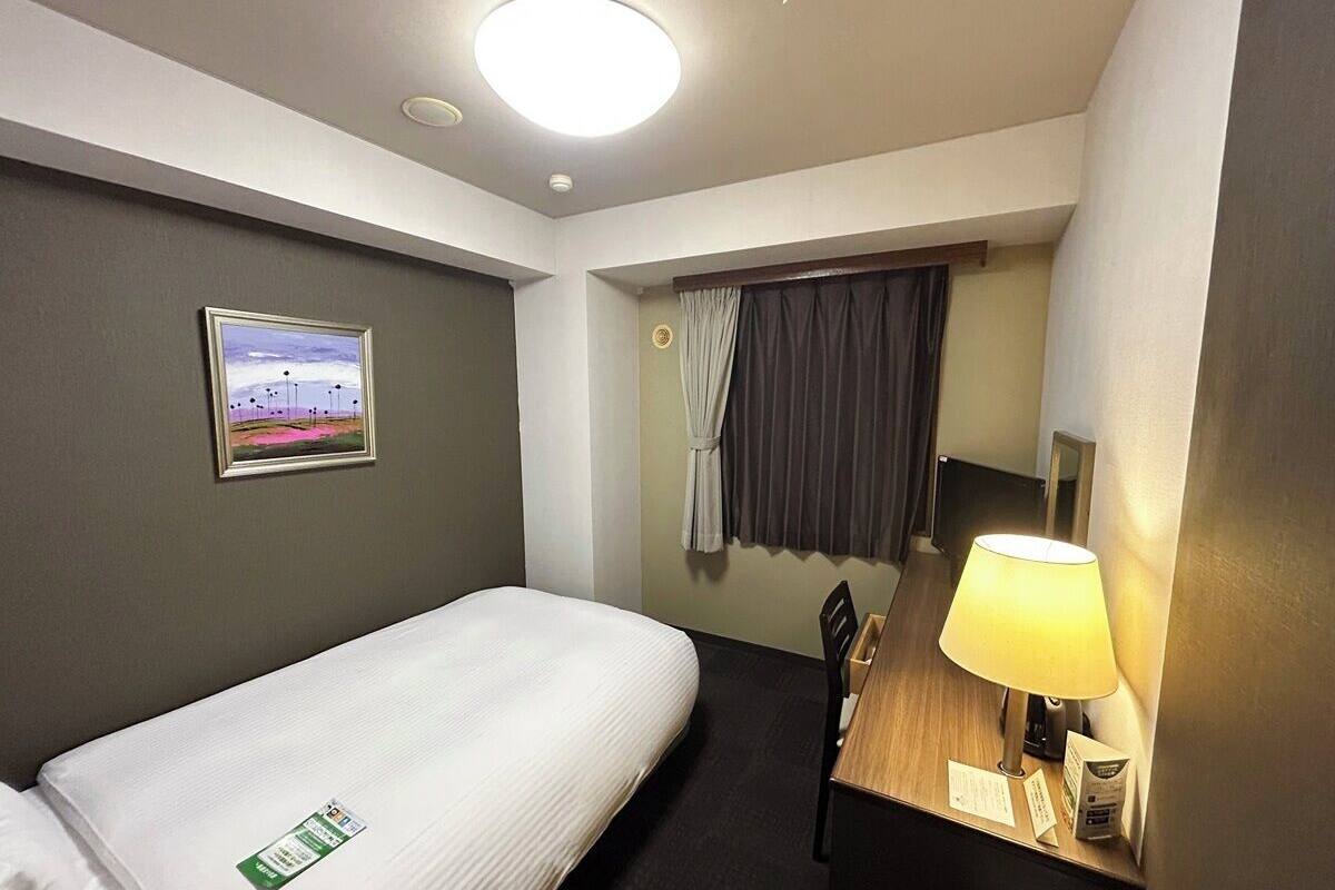 A hotel room with a single bed, a desk with a lamp, a chair, a framed picture on the wall, and a window with curtains.