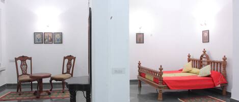 A room with a bed, two chairs, a small table, and a wall-mounted shelf.