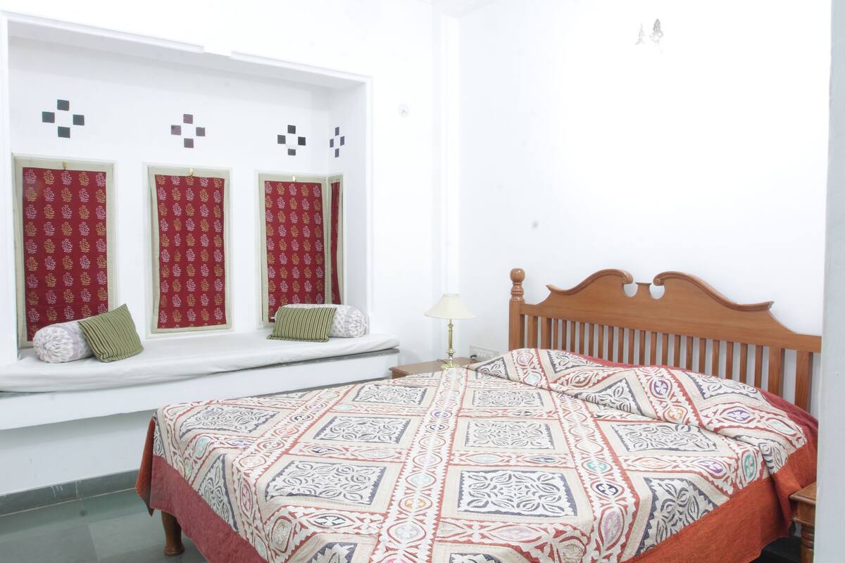 Un dormitorio con cama, un rincón con ventanal y cojines, y una mesita de noche.