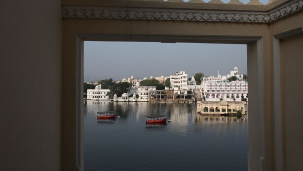 A view through an ornate doorway of a building with a water body in the foreground, featuring boats and a distant cityscape with traditional architecture.