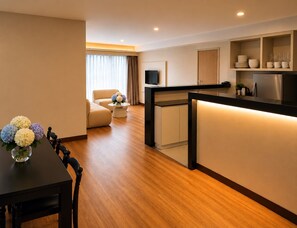 Une chambre d’hôtel moderne avec un plancher en bois, un coin repas comprenant une table et des chaises, un canapé, un coin cuisine avec un réfrigérateur et un téléviseur fixé au mur.