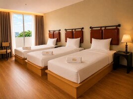 Habitación de hotel con tres camas, una silla, una mesita de noche con lámpara y vistas a zonas verdes.