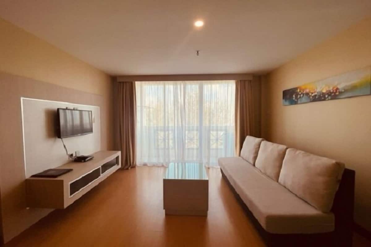 Un salon moderne avec un téléviseur à écran plat fixé au mur, un canapé beige, une table basse et une grande fenêtre ornée de rideaux légers.
