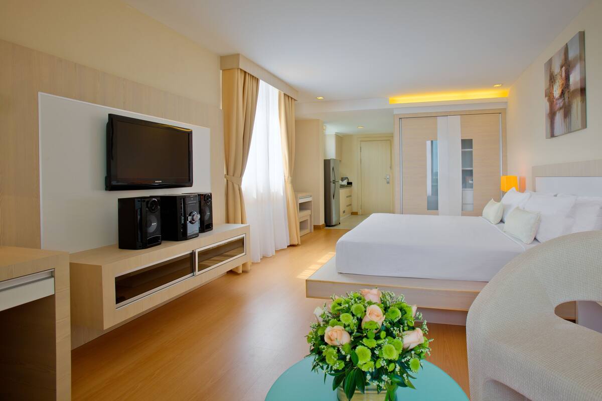 Une chambre d’hôtel comprenant un lit, une télévision, un bureau avec des haut-parleurs, un réfrigérateur, une table de chevet avec une lampe et un vase à fleurs.