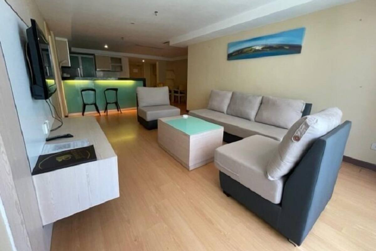 Un salon moderne avec un canapé, un pouf et une table basse. On aperçoit à l’arrière-plan un coin cuisine avec un comptoir-bar.
