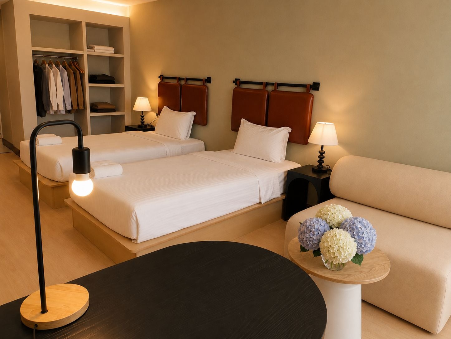 A hotel room with two beds, a sofa, a round table with a lamp, a bedside table with a lamp, and a wardrobe.