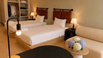 A hotel room with two beds, a sofa, a round table with a lamp, a bedside table with a lamp, and a wardrobe.