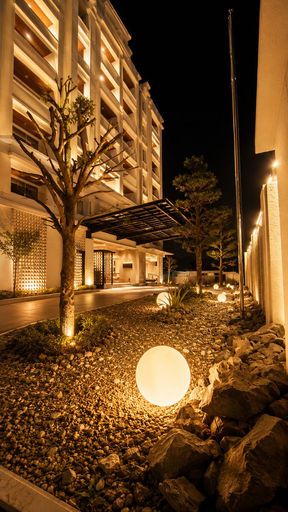A nighttime view of a building with a landscaped area featuring a tree, rocks, and a spherical light.