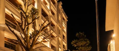 A nighttime view of a building with a landscaped area featuring a tree, rocks, and a spherical light.