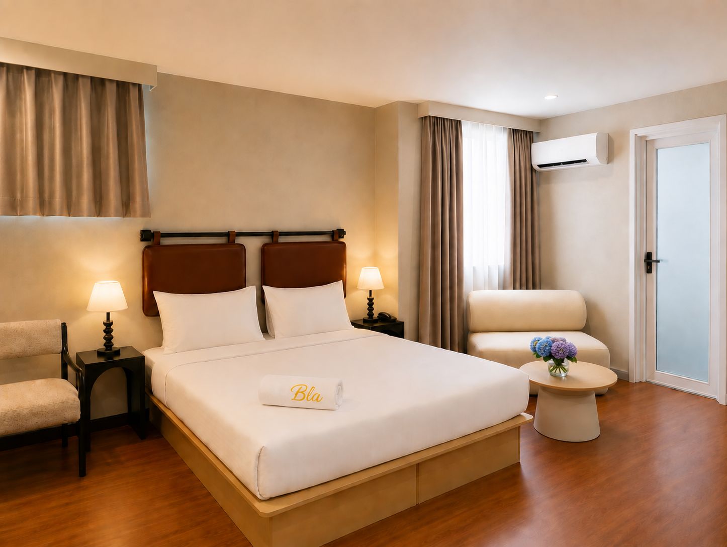 A hotel room with a bed, bedside tables, a chair, a round table, and an air conditioning unit.