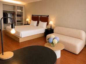 A hotel room with a bed, a sofa, a bedside table with a lamp, a nightstand, and a closet with hanging clothes. - Blanco Apartment Hotel (Bintulu)