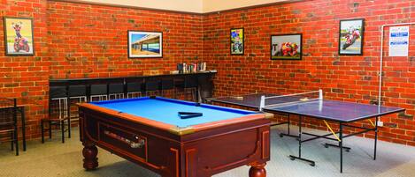 A billiards room with a pool table, a ping pong table, and a brick wall with framed pictures.