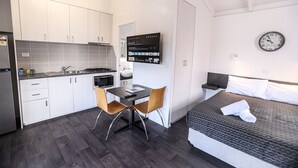 Een compacte hotelkamer met kitchenette, een bed met grijs gestructureerd matras, een wandklok en een flatscreen televisie waarop een nieuwskanal beeldschermt.