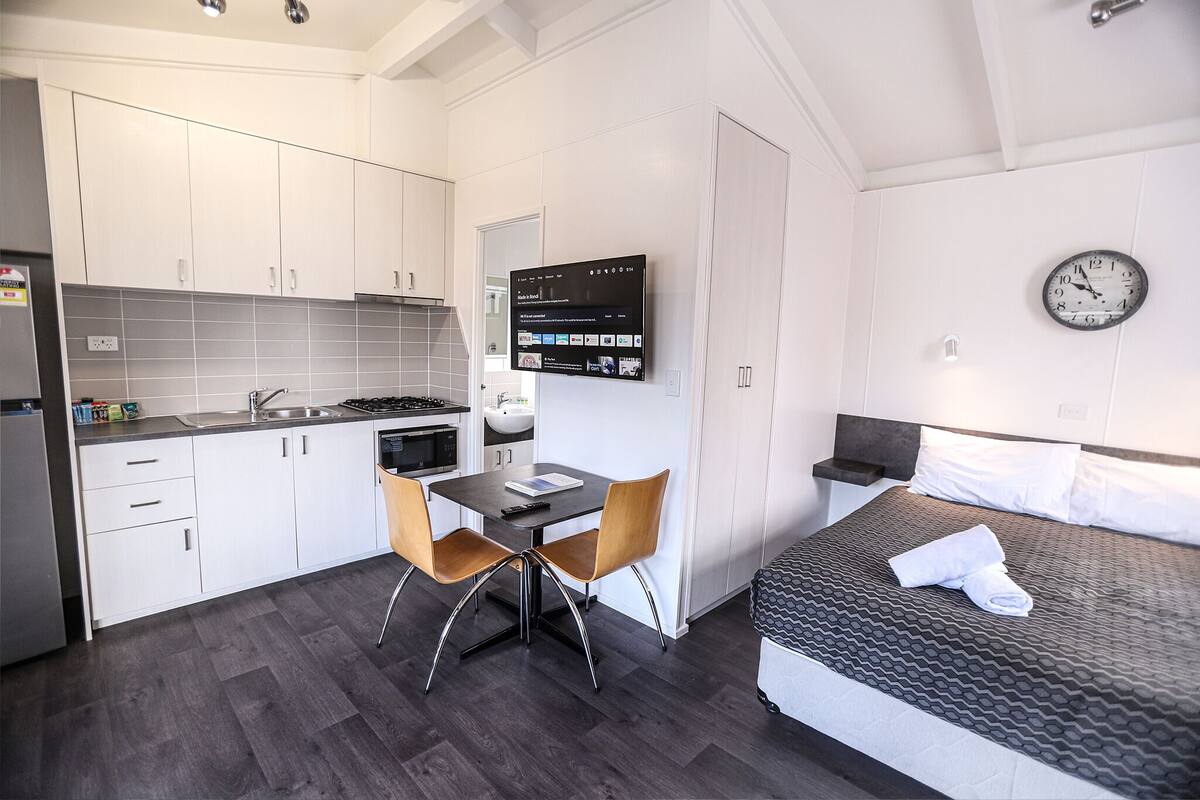 A compact hotel room with a kitchenette, a bed with a grey textured mattress, a wall-mounted clock, and a flat-screen TV displaying a news channel.