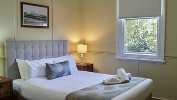 A neatly made bed with a grey headboard, white linens, and a grey patterned bedspread. There is a framed picture on the wall above the bed and a window with a view of trees.