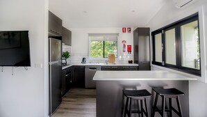 Een moderne keuken met een centraal eiland, barstoelen, een inbouwoven en een raam met uitzicht op groen.