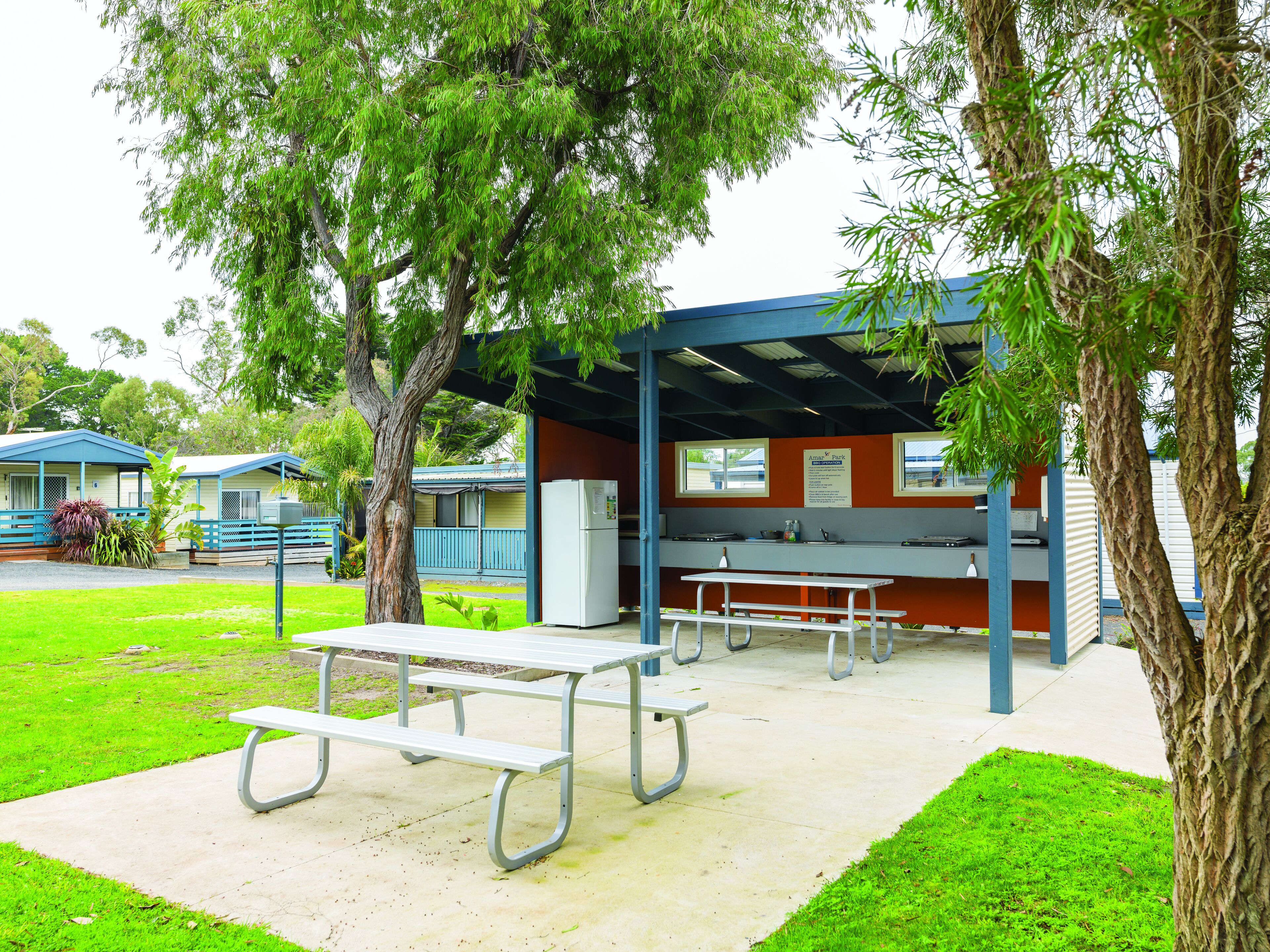 Zona de picnic con mesas y un área de estar cubierta, rodeada de árboles y un edificio con un cartel.