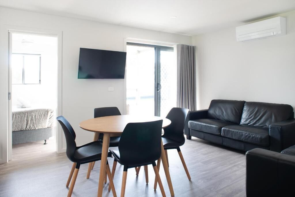 A modern living room with a round wooden table, black chairs, a black leather sofa, and a flat-screen TV mounted on the wall.