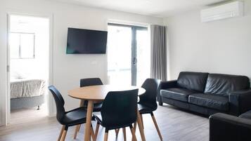 A modern living room with a round wooden table, black chairs, a black leather sofa, and a flat-screen TV mounted on the wall.